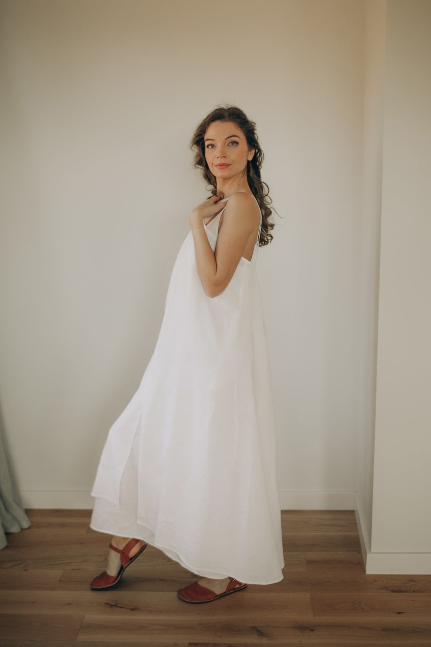 Woman wearing a white linen dress standing in a room with wooden floor and neutral walls.