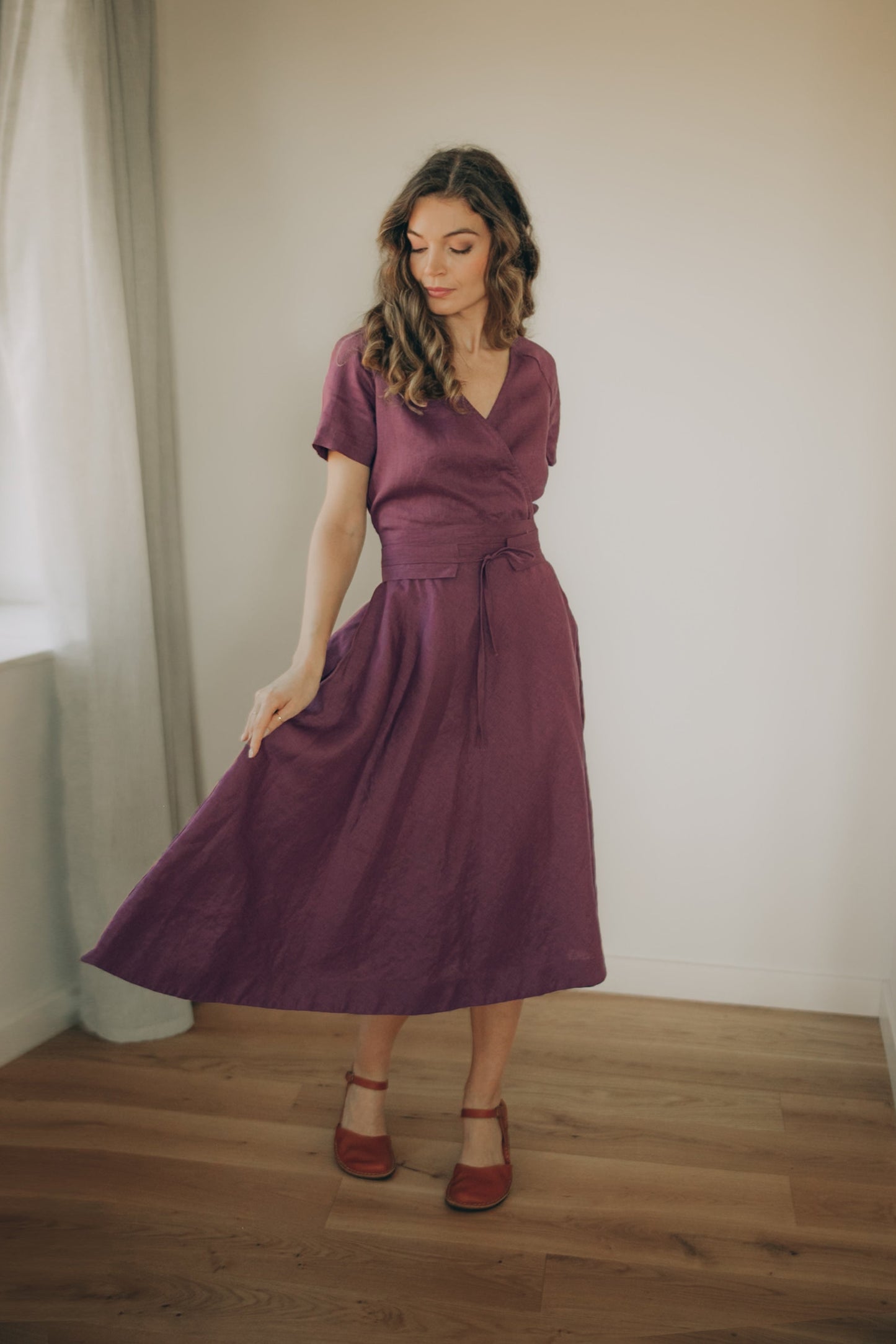 Woman wearing a purple linen midi dress standing in a room with wooden floor and white walls.