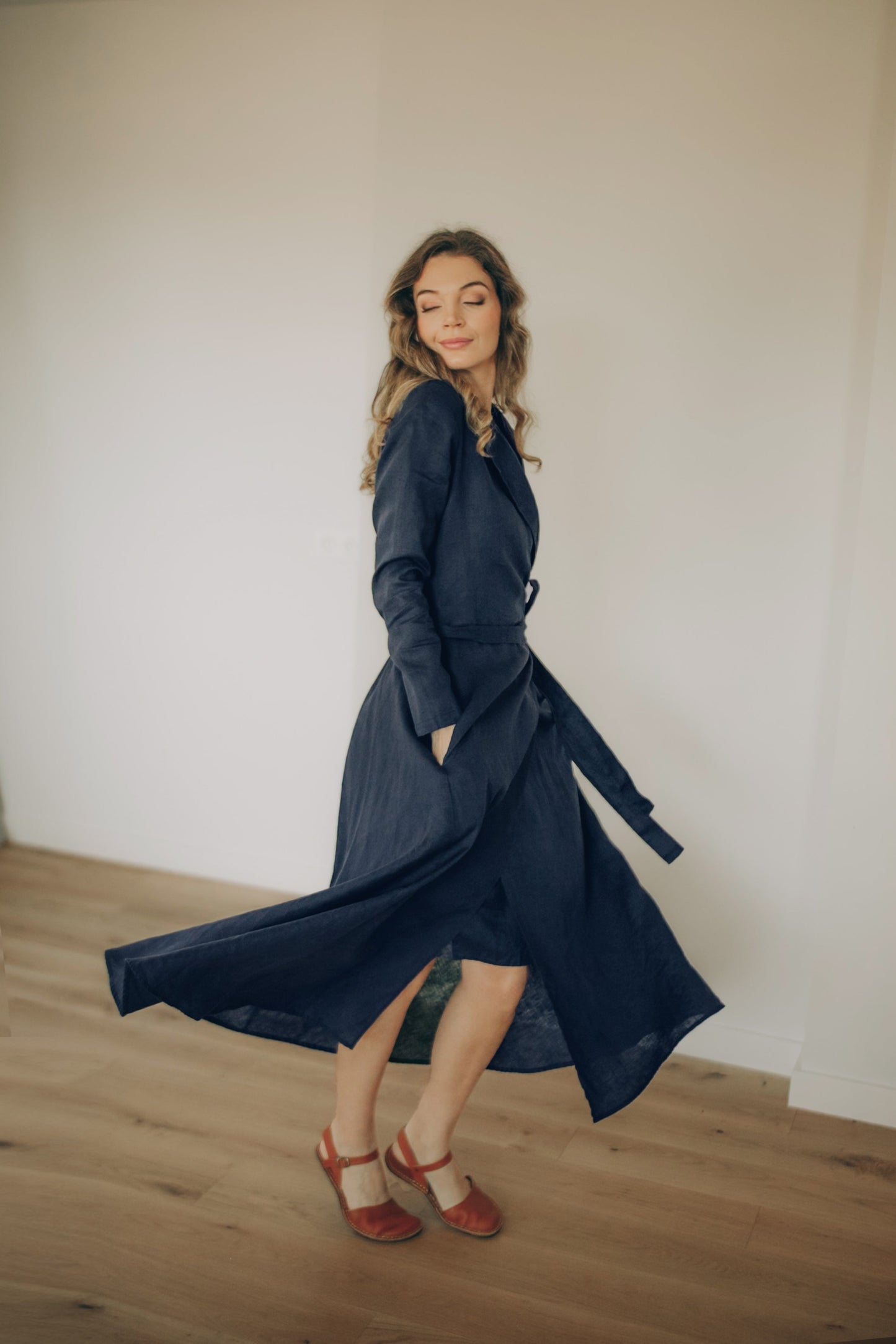 Woman wearing a navy blue linen wrap dress with a belt, standing against a plain white wall.