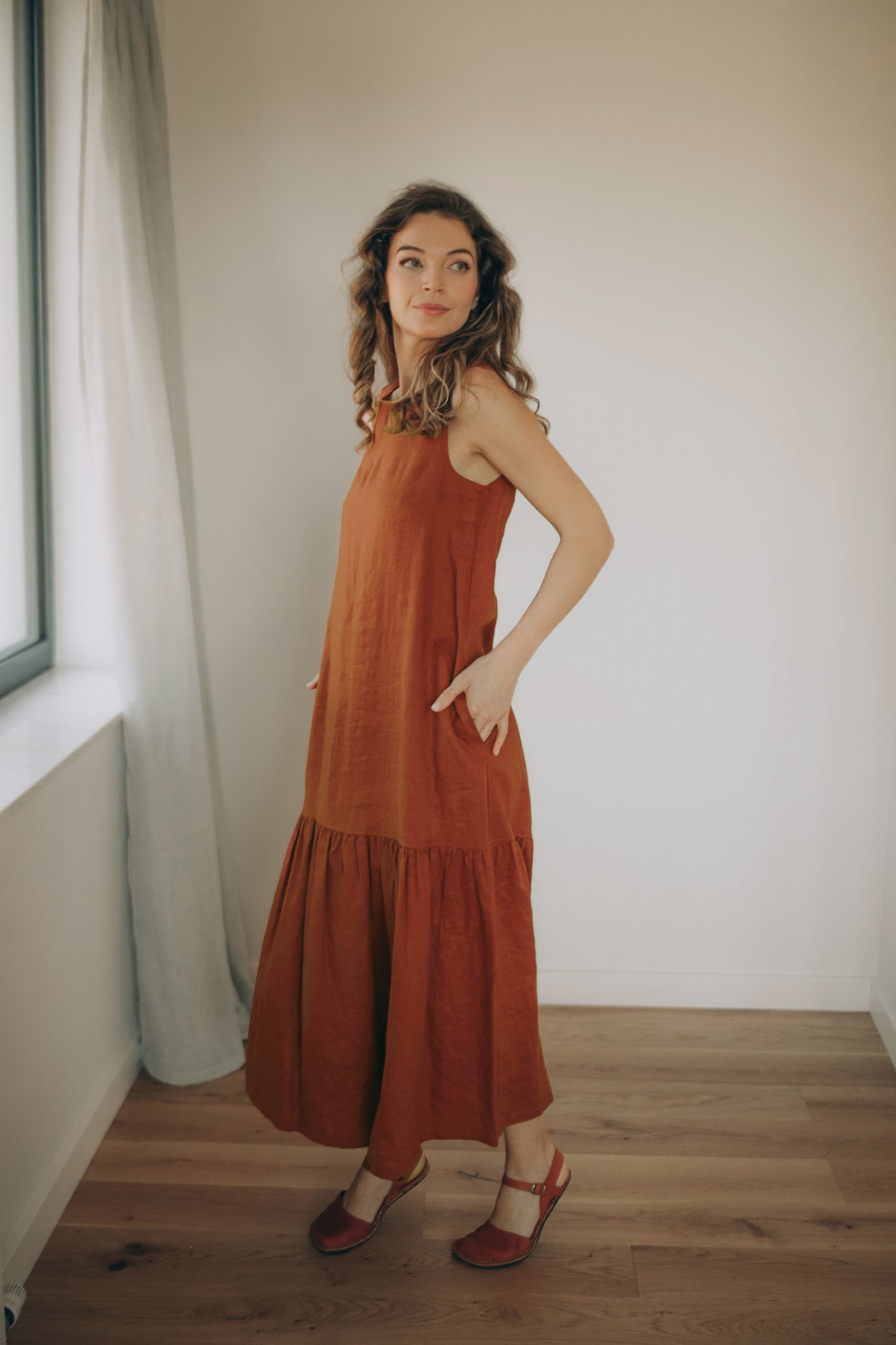 Woman wearing a rust-colored linen dress standing in a room with wooden flooring and a window.