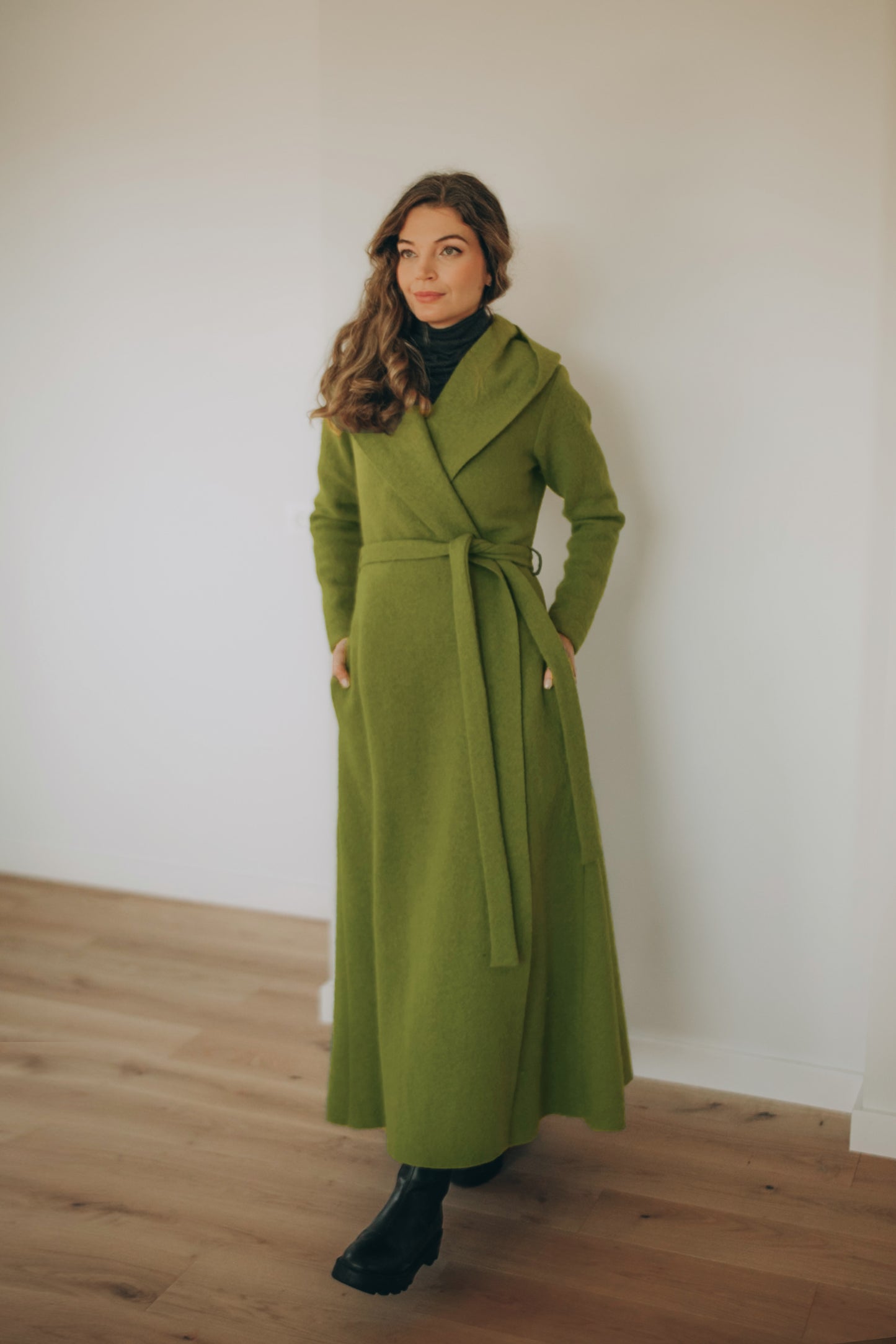 Woman wearing a long green wool coat standing against a white wall.